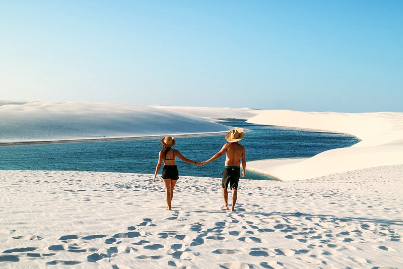 Casal de mãos dadas caminhando nas dunas brancas em direção à lagoa azul